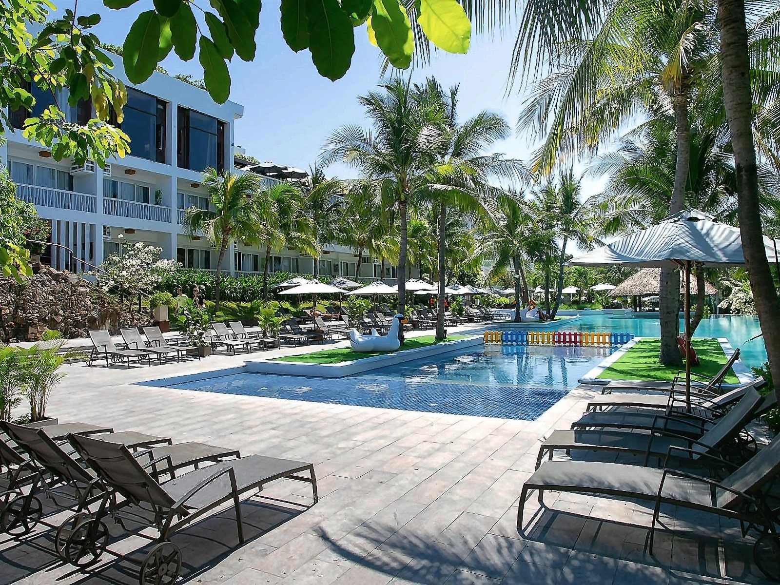 Long swimming pool lined with sun loungers and umbrellas below white resort buildings