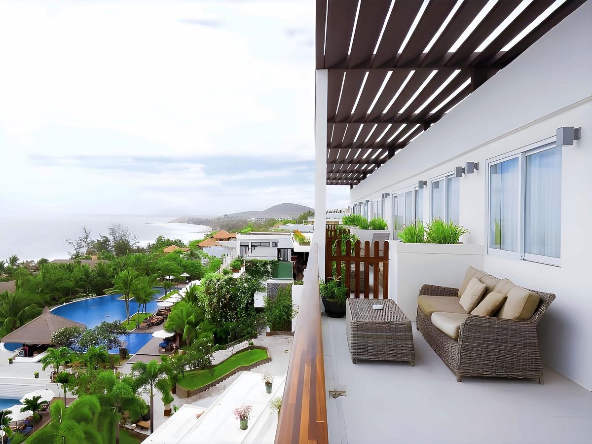 Balcony with wicker sofa looking over gardens and pools to the sea and a rocky headland