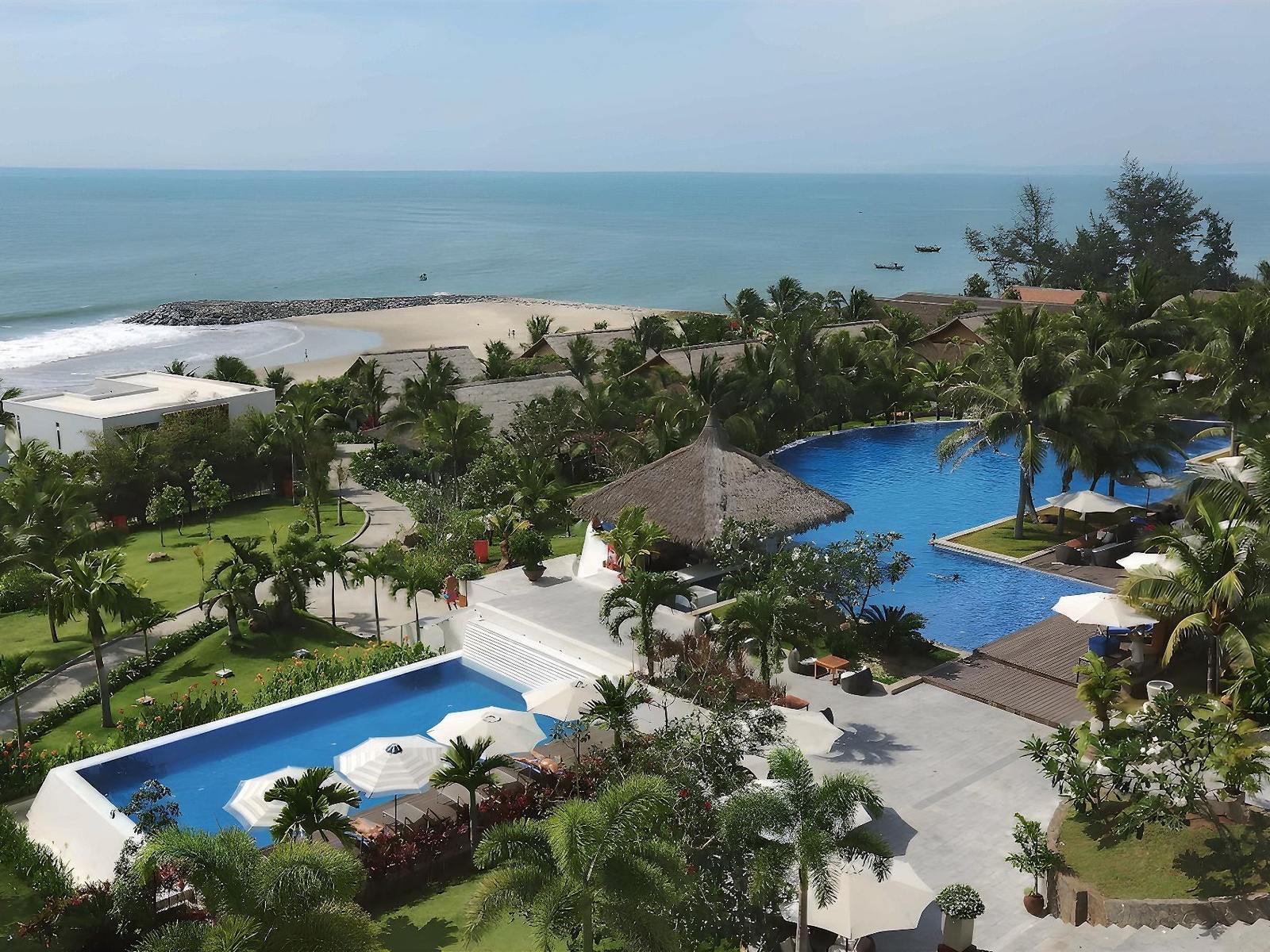 Aerial view of pools, palm gardens and white resort blocks beside a sandy beach and blue sea